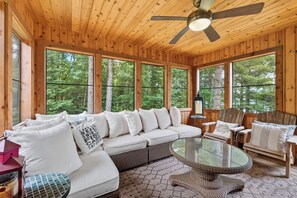 Interior de una cabaña de madera con sofá, sillas, una mesa de vidrio y ventanas amplias.