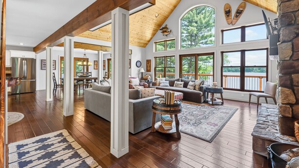 A spacious living area with wooden beams, large windows, a stone fireplace, and a cozy seating arrangement. - Gliemwood II at Clam Lake | 5BR Lakefront Retreat (Clam Lake)