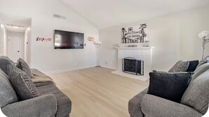 Uma sala de estar com lareira, uma TV de tela plana na parede e dois sofás cinzas com almofadas estampadas.
