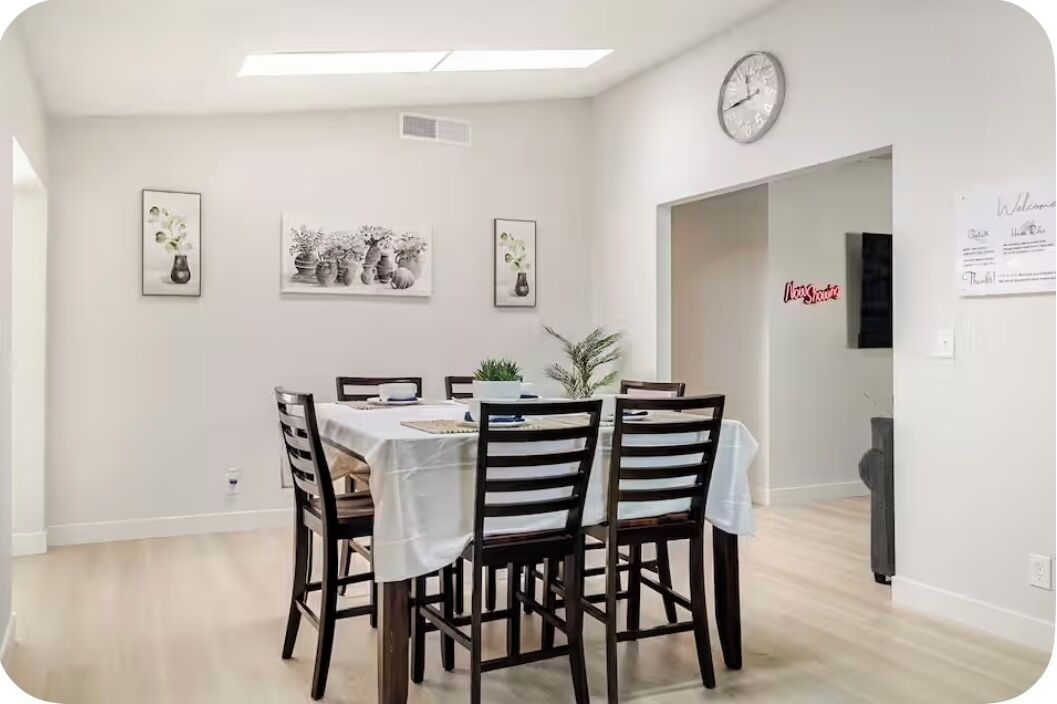 Ein Essbereich mit einem Tisch, gedeckt für vier Personen, umgeben von Stühlen. An der Wand hängen gerahmte Bilder, eine Uhr und ein Schild mit der Aufschrift 'Welcome'.