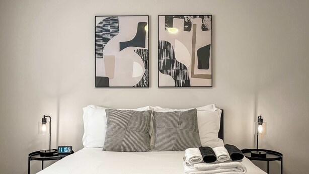A neatly made bed with a white comforter and two pillows, flanked by two bedside tables with lamps and clocks. - Chic Skokie Retreat - 1BR Unit in Skokie (Skokie)