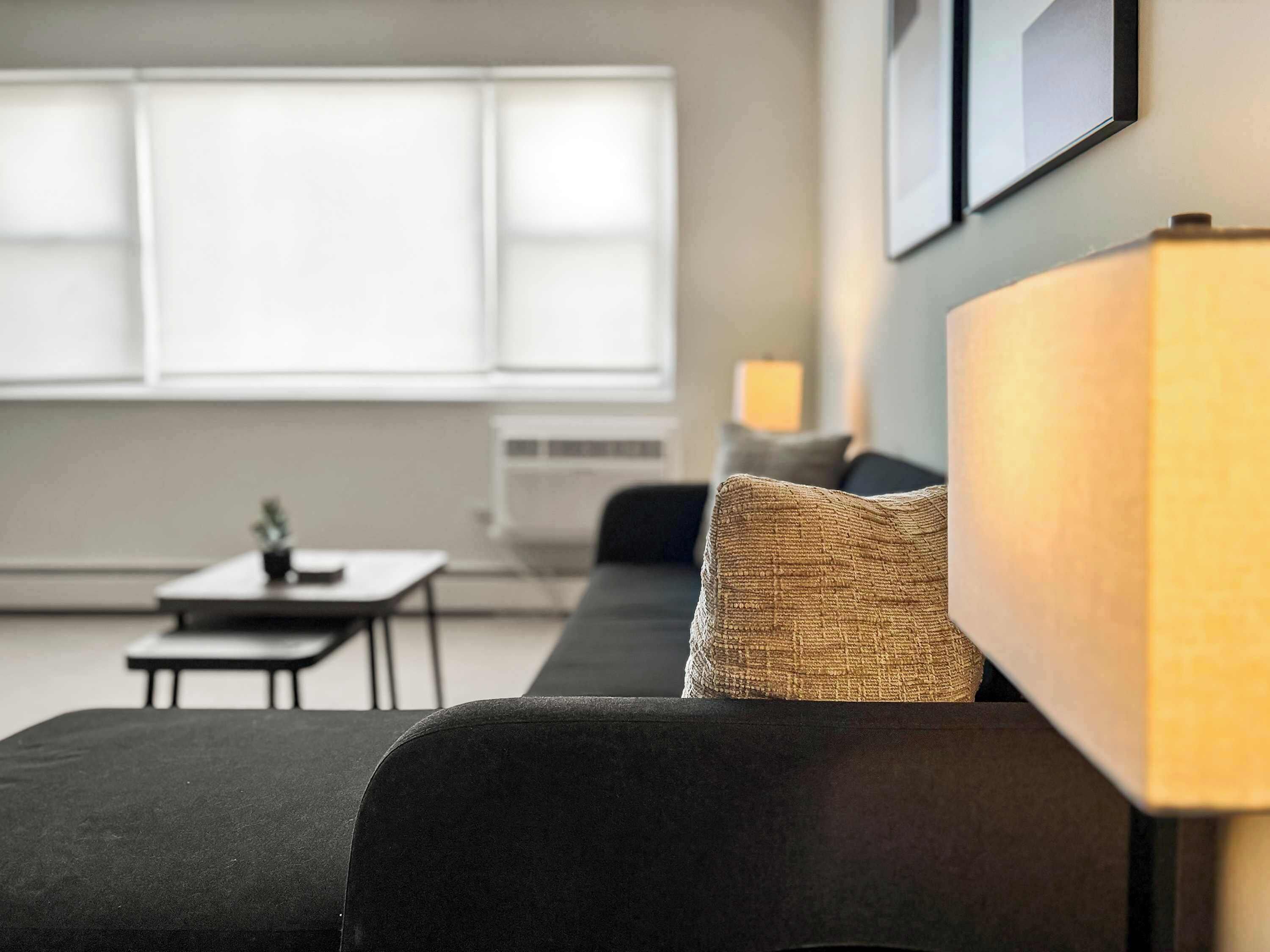 A modern living room with a dark sofa, a small table, and a lamp. There is a window with blinds and a wall-mounted picture frame.