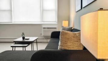 A modern living room with a dark sofa, a small table, and a lamp. There is a window with blinds and a wall-mounted picture frame.