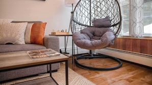 A cozy living room with a grey sofa, a round hanging chair, a small table with a magazine, and a chessboard.
