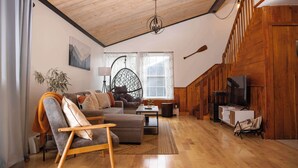 A cozy living room with a wooden ceiling, a large window with curtains, a sofa, a chair, and a television.