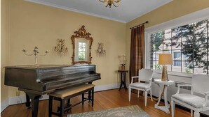 A room with a grand piano, a chandelier, a large mirror, and a view of trees through a window. - 5 Bedroom Home w/ Private Parking Near Niagara Falls (Niagara Falls)