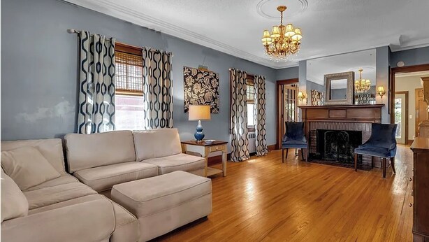 A living room with a beige sofa, a blue armchair, a wooden fireplace, and a chandelier. - 5 Bedroom Home w/ Private Parking Near Niagara Falls (Niagara Falls)