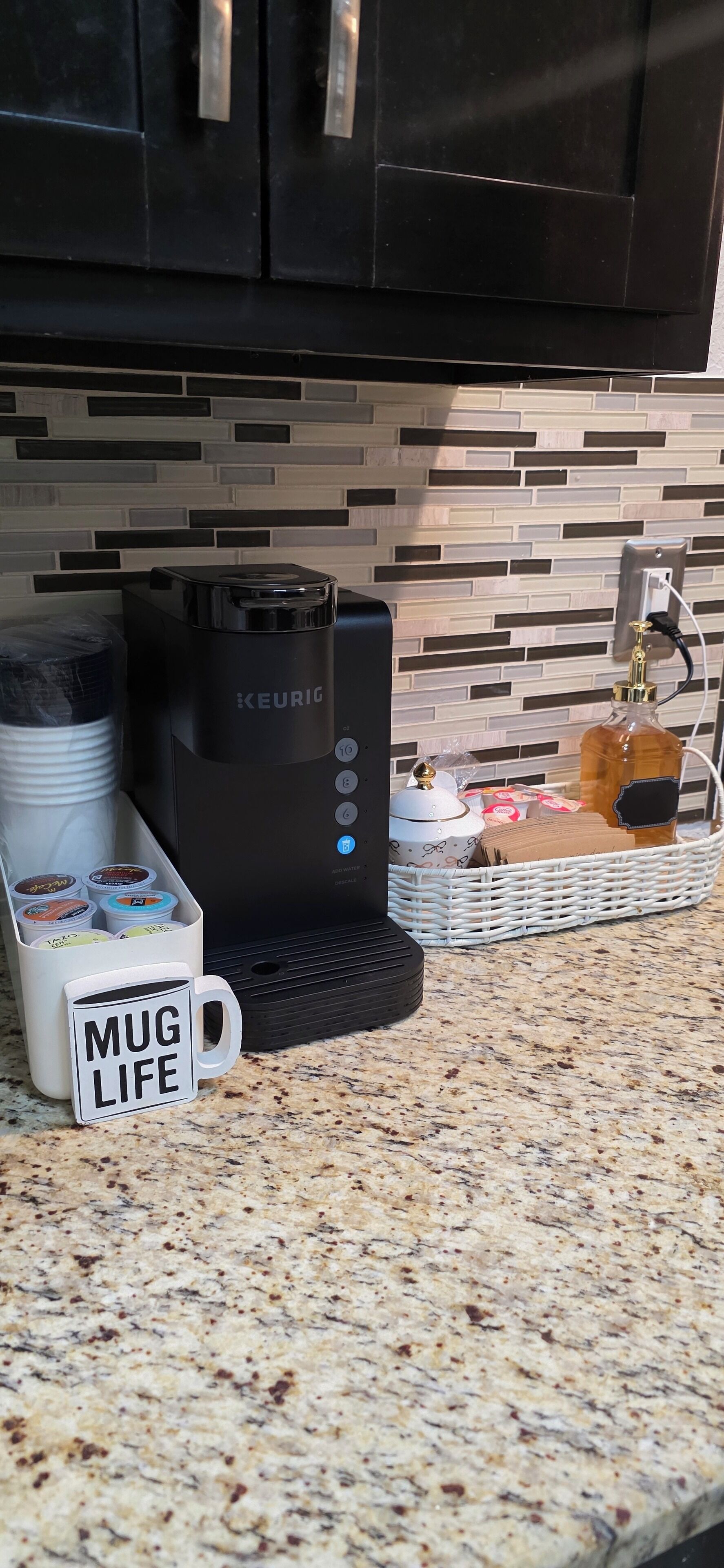 Una cocina con cafetera Keurig, una taza con la etiqueta 'Mug Life', una cesta con diversos objetos y un salpicadero de azulejos.