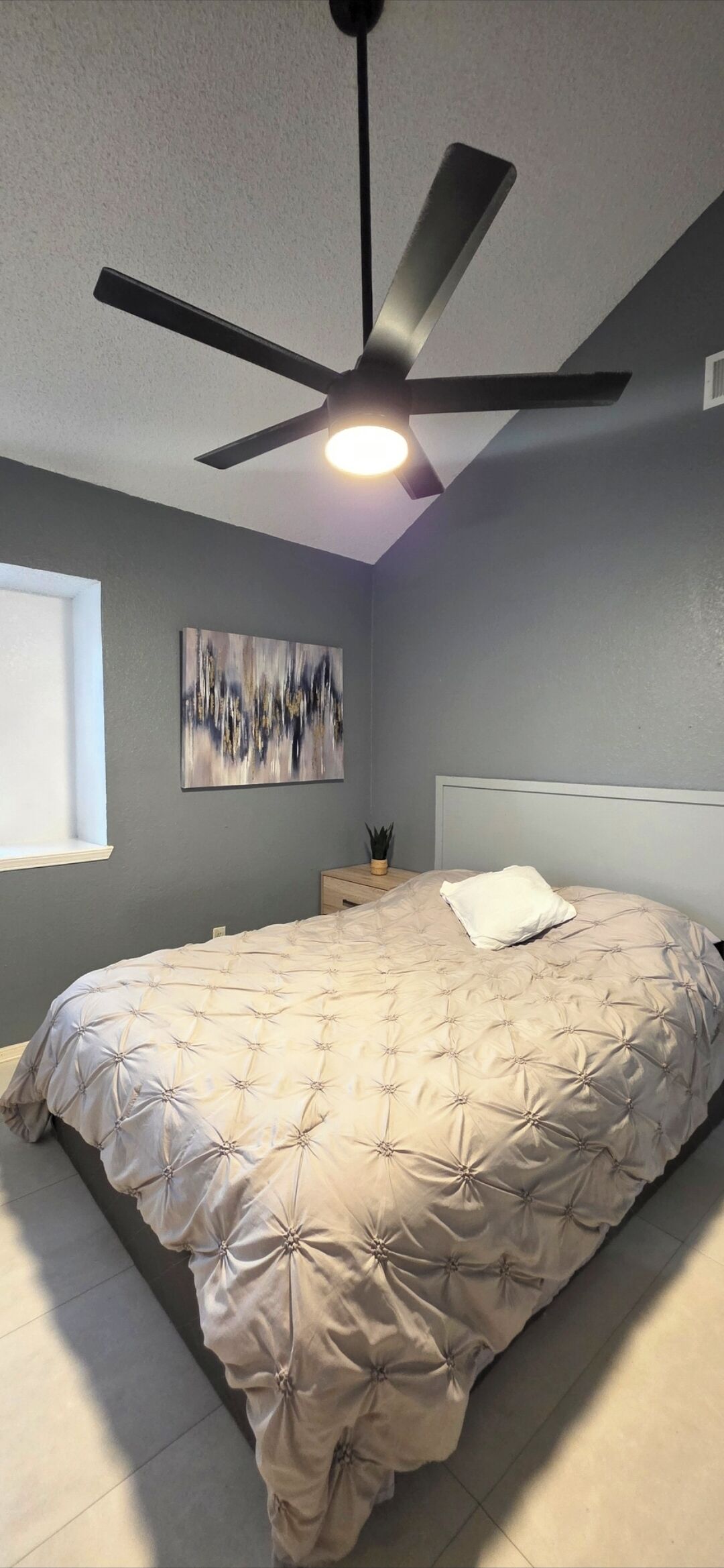 Une chambre à coucher comprenant un lit, un ventilateur de plafond, une œuvre d’art accrochée au mur et une table de chevet avec une lampe.