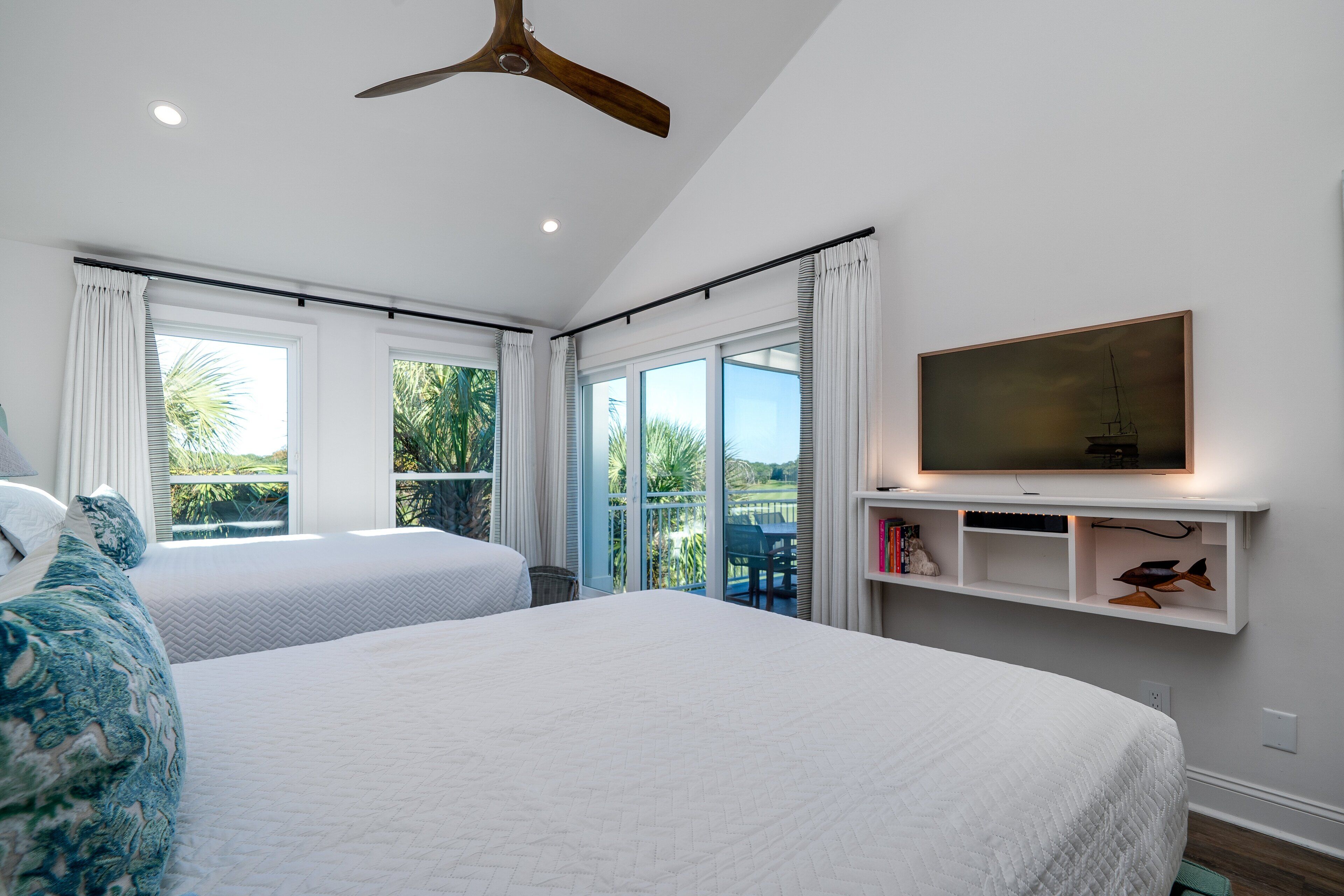 A bedroom with two beds, a ceiling fan, a TV on a shelf, and a balcony with a view of greenery.