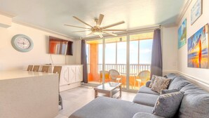 Un vaste salon avec un canapé gris, une petite table et un ventilateur de plafond. Il y a un balcon avec vue sur le ciel et un coin repas avec des chaises.