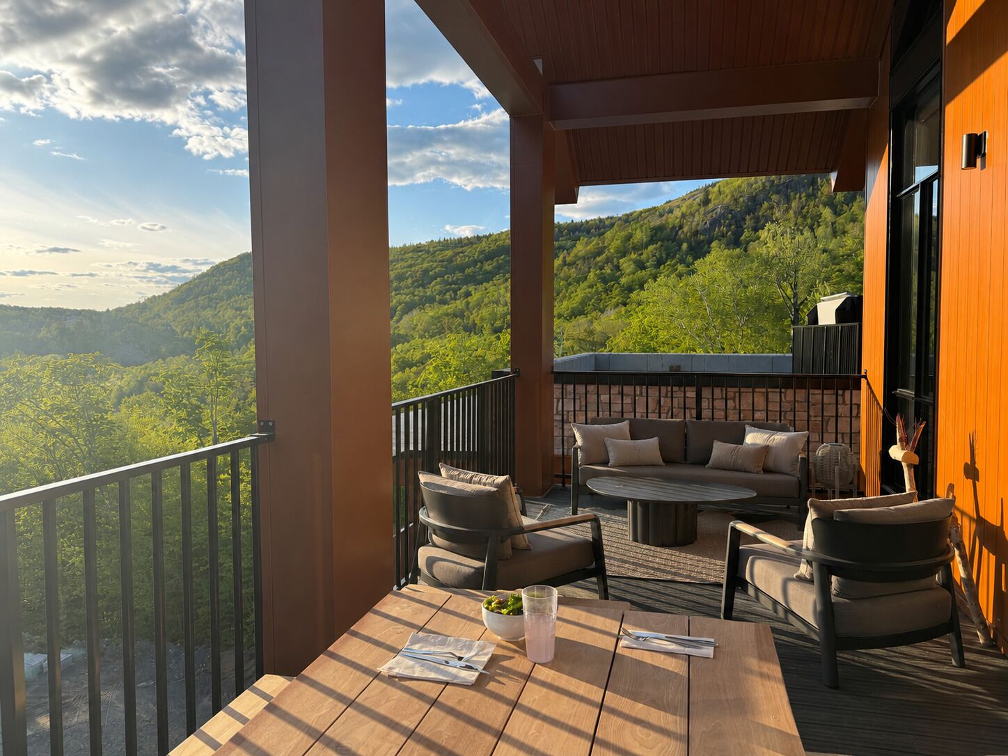 A balcony with outdoor furniture, a table with a drink, and a view of a forested mountain.