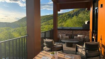 A balcony with outdoor furniture, a table with a drink, and a view of a forested mountain.