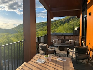 A balcony with outdoor furniture, a table with a drink, and a view of a forested mountain.
