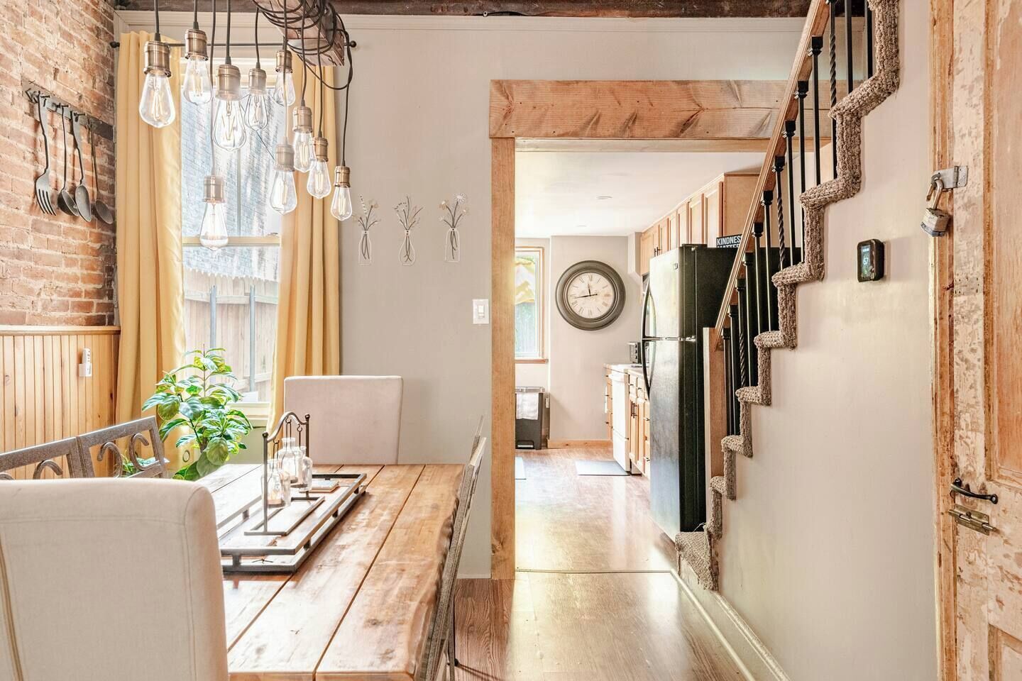 A rustic kitchen with exposed brick walls, wooden beams, a wooden dining table with chairs, and a staircase with a woven railing.