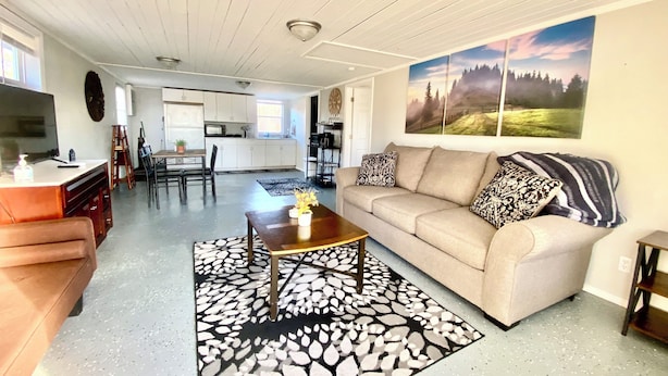 A spacious living room with a sofa, coffee table, and patterned rug. A kitchen is visible in the background with white cabinets and a dining area.