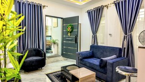 Un salon moderne avec un canapé bleu, un fauteuil noir et une table basse. Une porte ornée d’une guirlande décorative et un ventilateur se trouvent sur le côté droit.