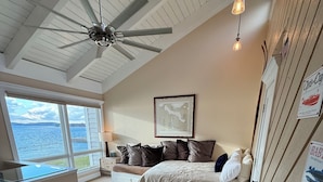 Une chambre avec une grande fenêtre, un ventilateur de plafond, un lit avec des oreillers et un mur orné de tableaux.