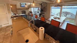 Une cuisine dotée d’un grand îlot central, d’un évier et offrant une vue sur l’eau. On y trouve des chaises et une table, et un escalier est visible au fond.