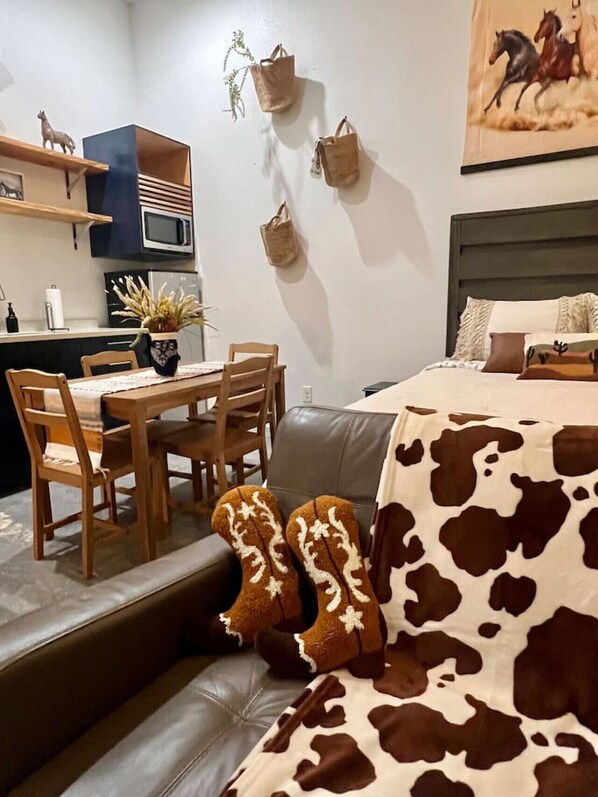 Een moderne woonkamer met een leren bank, een eettafel met stoelen, een keuken met een magnetron en kastjes, en een bed met een deken met koeienprint.