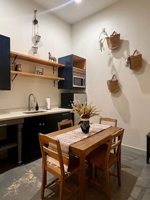 Een kleine keuken met houten kastjes, een magnetron en een spoelbak. Er staat een eettafel met stoelen en een vaas met gedroogde planten.