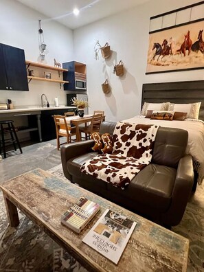 Een moderne woonkamer met een leren bank, een salontafel met tijdschriften, een eethoek met een houten tafel en stoelen, en een keuken met donkere kastjes en een magnetron.