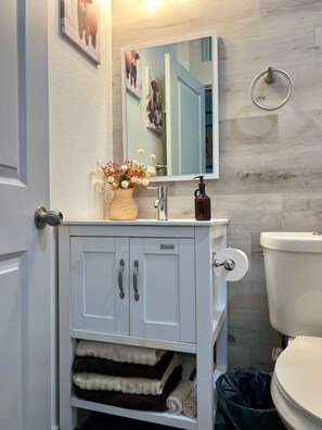 Een badkamer met een wit wastafelmeubel, een spiegel, een toilet en een prullenbak.