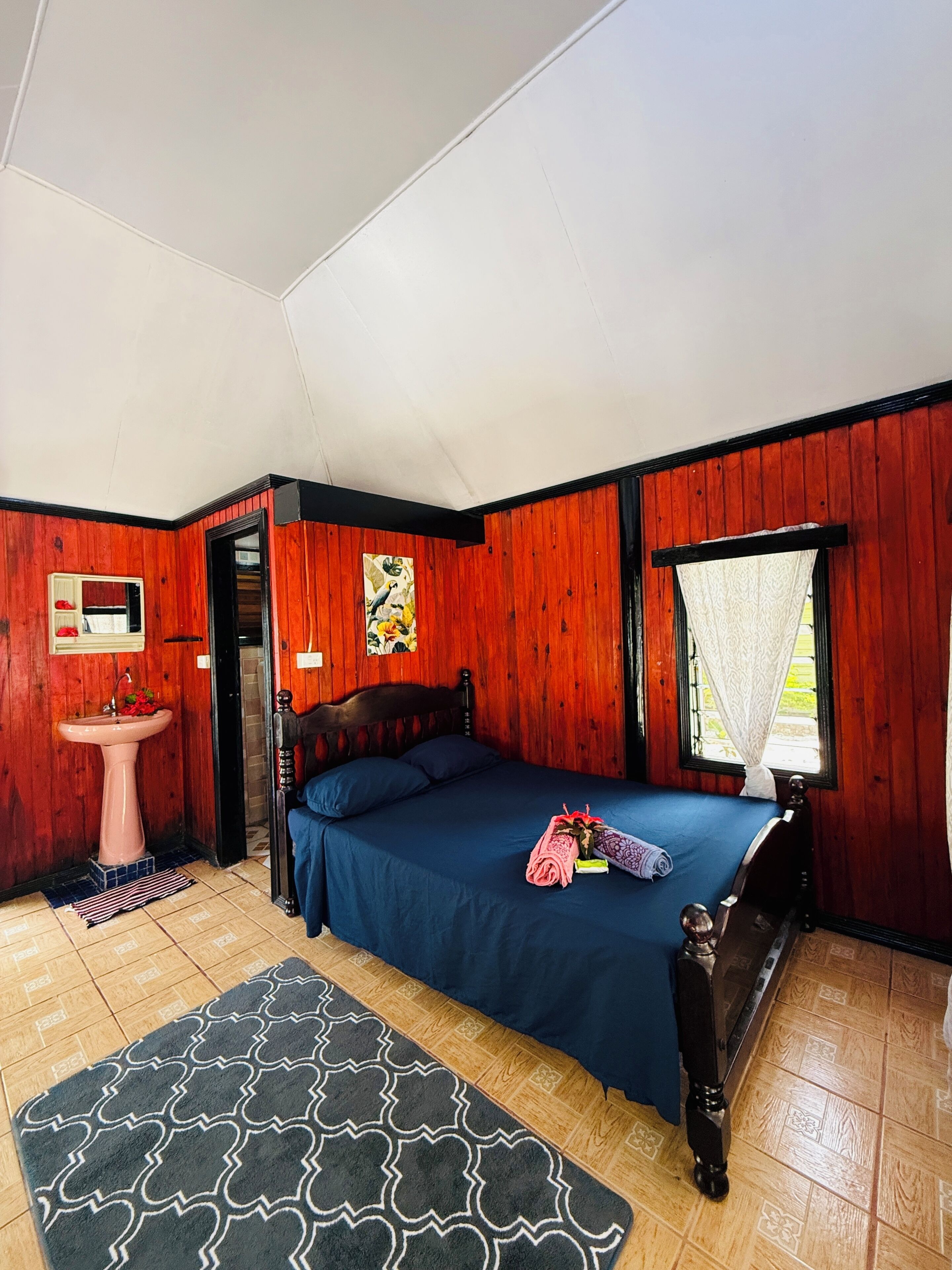 A room with a bed, a wooden wall, a window with white curtains, a sink, and a door leading to another room.