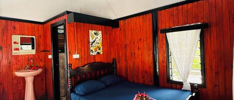 A room with a bed, a wooden wall, a window with white curtains, a sink, and a door leading to another room.