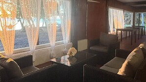 Una habitación con mobiliario de mimbre, una mesa de centro de vidrio y ventanas amplias con cortinas translúcidas.