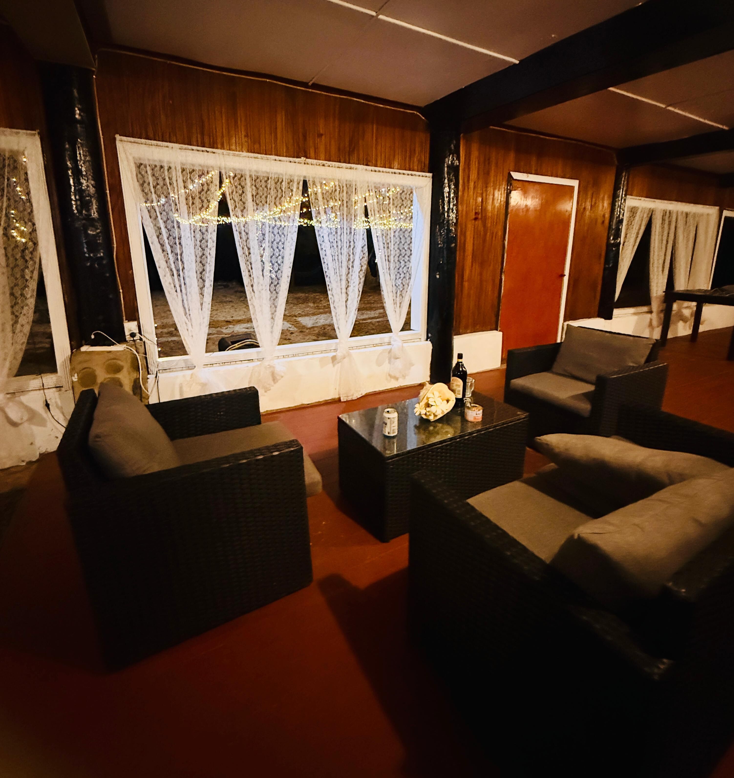 Een gezellige woonkamer met rieten meubels, een bijzettafeltje en een groot raam met kantgordijnen.