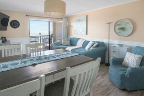Et spiseområde med et træbord, hvide stole og en blå servietring. Der er en teal sofa med mønstrede puder og en gulvlampe.