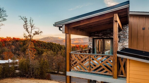 Una baita di legno con un balcone affacciata su una zona boschiva e montagne.