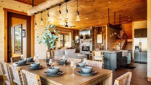 Un'ampia sala da pranzo con un tavolo in legno apparecchiato per sei persone, circondato da poltrone imbottite. Sullo sfondo, si nota un camino in pietra e una cucina con elettrodomestici moderni.