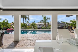 Uma área de refeições à beira da piscina, com vista para uma piscina, palmeiras e um edifício ao fundo.
