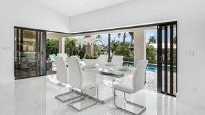 A modern dining area with a glass table and white chairs, large windows, and a view of a pool and greenery. - Sunshine, Serenity & a Heated Pool Escape for All-Day Fun – Modern Vibes Villa - Roelens (Cape Coral)