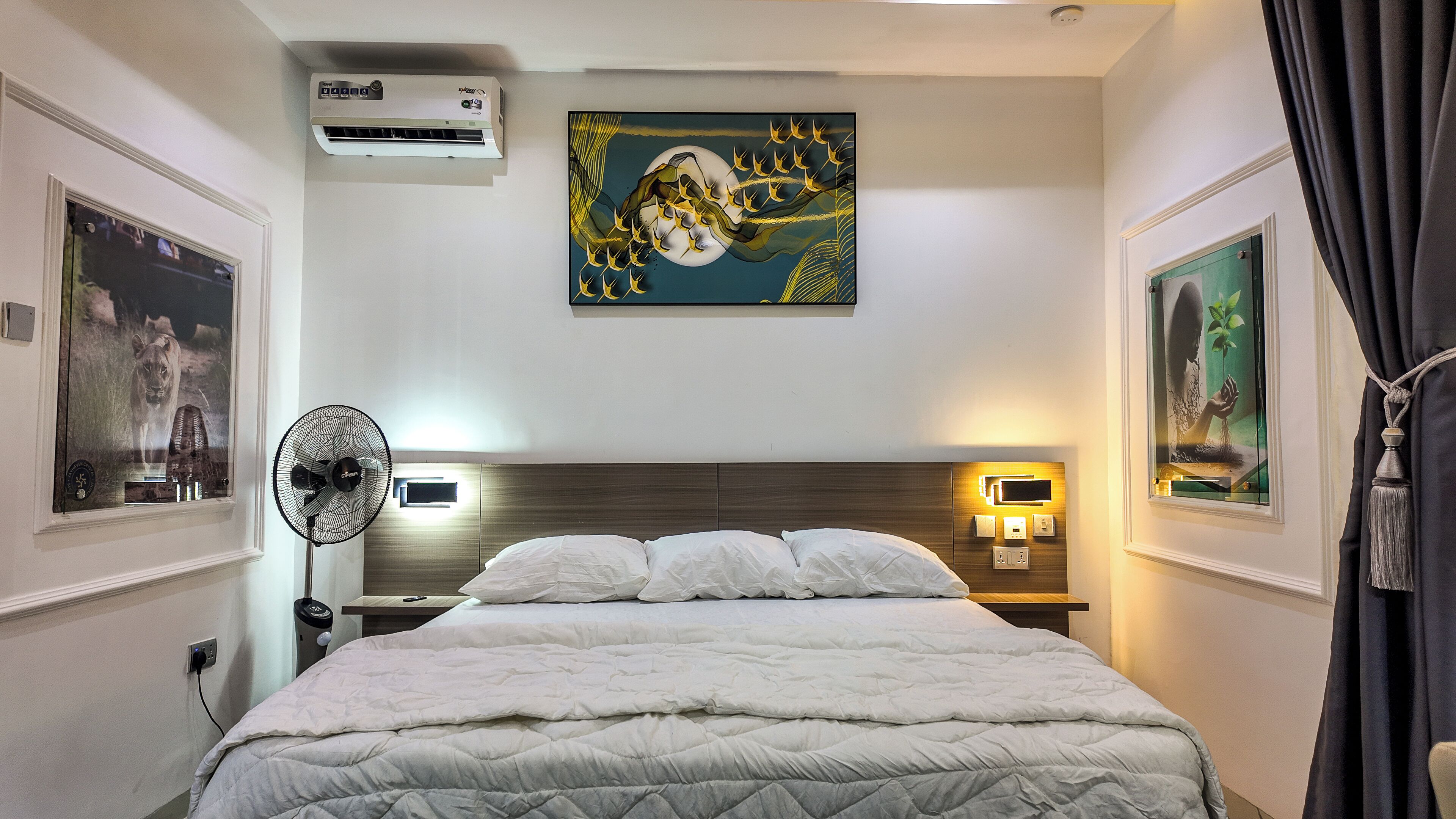 Habitación de hotel con cama, aire acondicionado, ventilador de pared, un cuadro y mesita de noche con lámpara.