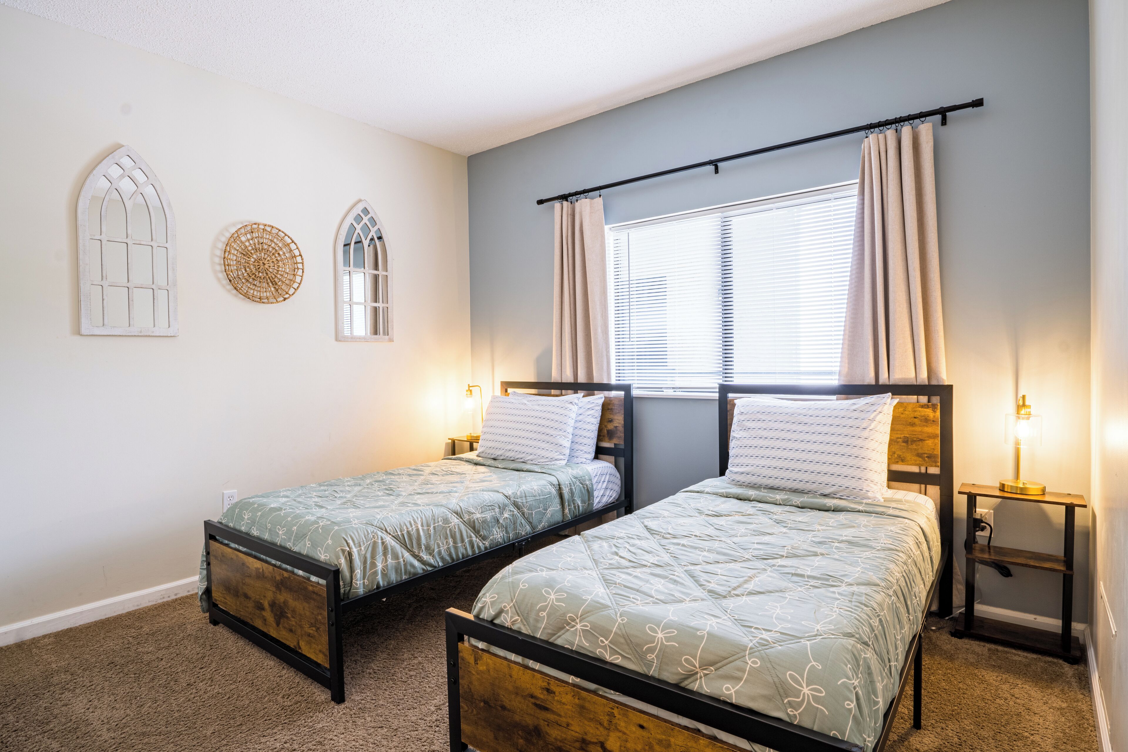 A bedroom with two single beds, a window with blinds, a wall-mounted shelf, and decorative wall art.