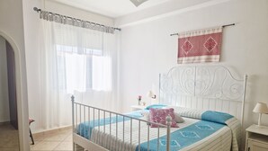 Un dormitorio con estructura de cama metálica blanca, colcha estampada en azul y blanco, cabecera decorativa roja y blanca, y una mesita de noche con lámpara.