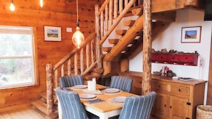 Une salle à manger en bois avec une table dressée pour quatre personnes, des chaises à rayures, un escalier en bois et un buffet rouge rempli de livres.