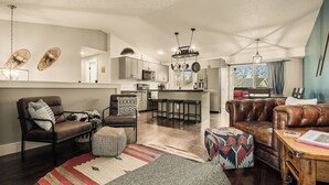 A modern living room with a sofa, ottoman, and coffee table. A kitchen with a bar and stools is visible in the background. - Family Arcade Retreat + Fire Pit + 4BR Near RMNP (Loveland)