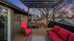 A covered outdoor deck with a wooden floor, a red sofa, a red chair, and a table. - Family Arcade Retreat + Fire Pit + 4BR Near RMNP (Loveland)
