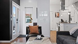 A modern living room with a black leather sofa, a marble coffee table, and a black leather armchair. There is a kitchen area with white cabinets and a stainless steel range hood. - Urban Retreat @ Cherry Street (Tulsa)