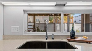 A modern kitchen with a large sink, a window with a view of the neighborhood, and a countertop with a bottle and brush. - Urban Retreat @ Cherry Street (Tulsa)