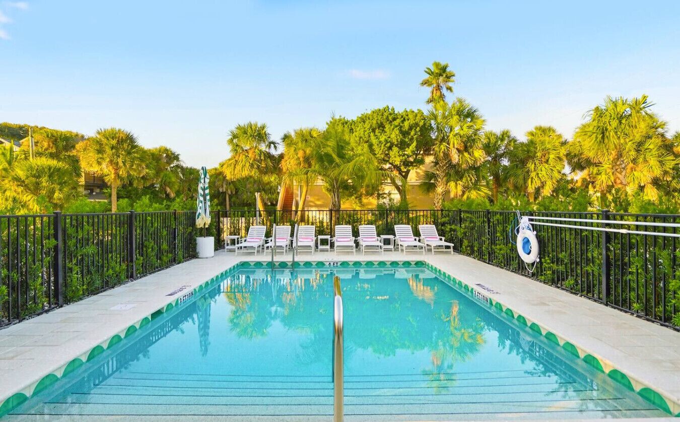 Una piscina con lettini e un galleggiante, circondata da palme e da una recinzione.