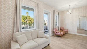 Ein geräumiges Wohnzimmer mit einem beigen Sofa, zwei rosa Sesseln und einem Holztisch. Große Fenster bieten Blick auf Palmen und einen Balkon.