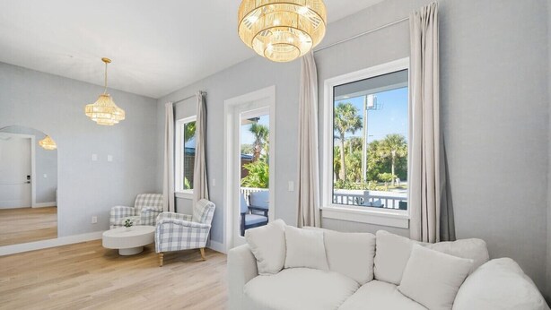 Una sala de estar moderna con un sofá blanco, un sillón a cuadros y una mesa redonda de centro. Hay dos ventanas grandes con cortinas, y un candelabro cuelga del techo.