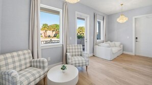 Un salón espacioso con un ventanal grande, un sofá blanco, un sofá a cuadros, una mesa de centro redonda y un balcón con vistas a palmeras.