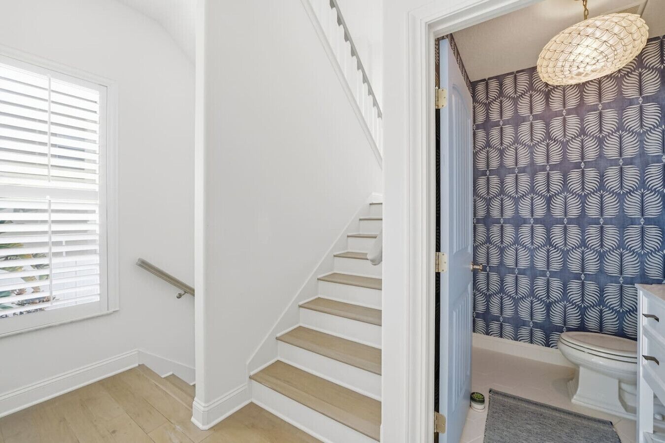 A staircase leading to a bathroom with patterned wallpaper, a hanging light fixture, and a window with blinds.