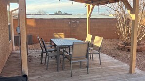 Un patio al aire libre con una mesa y sillas de madera, una parrilla y vistas a un muro de ladrillo y árboles.